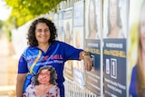 A woman with curly dark hair, smiling and leaning against a fence with election corflutes on it.