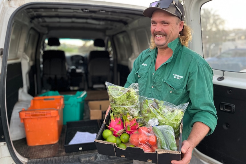 man smiles next to open van holding a box of vegetables