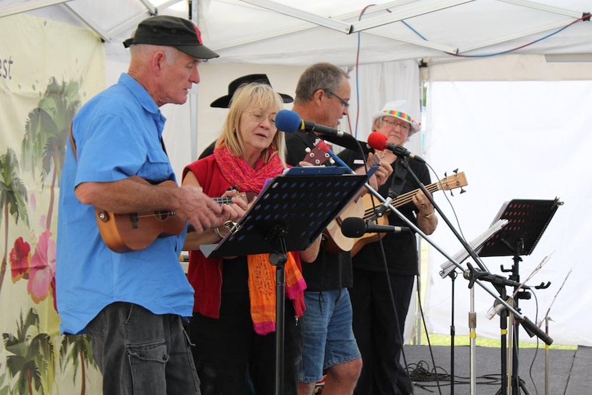 Five mature aged people on stage playing ukuleles and reading music with microphone out front.