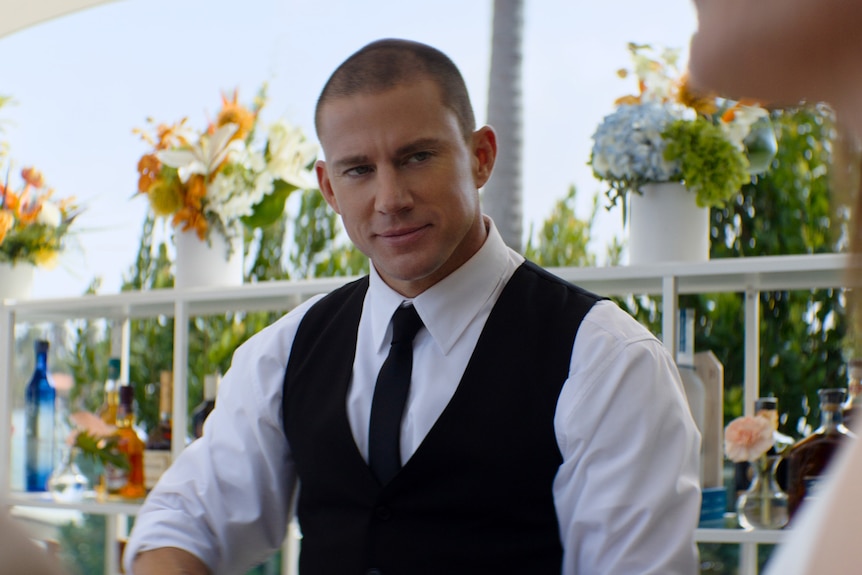 A 40-something man with a buzzcut, dressed smartly in a vest and tie, smiles slightly, while standing behind a bar