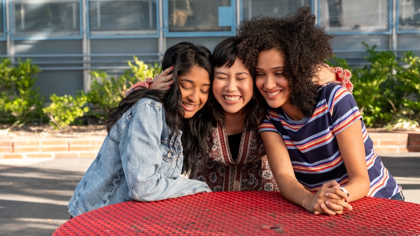 Three teenage girls sit and hug at a red table outside