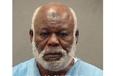 A bald man with a short silver beard poses for a mugshot while in custody in a light blue top.