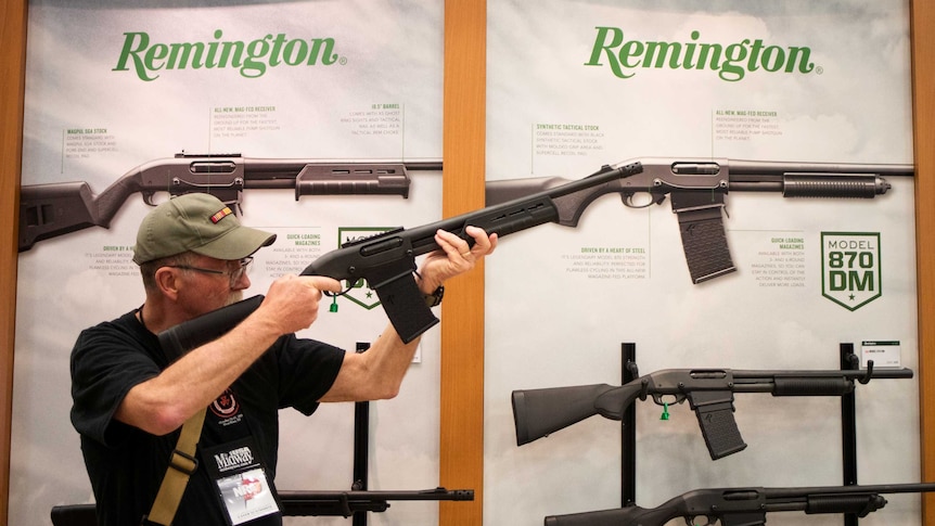 A man aims a firearm while standing in front of a Remington branded poster with display model guns.