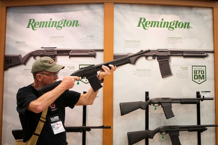 A man aims a firearm while standing in front of a Remington branded poster with display model guns.