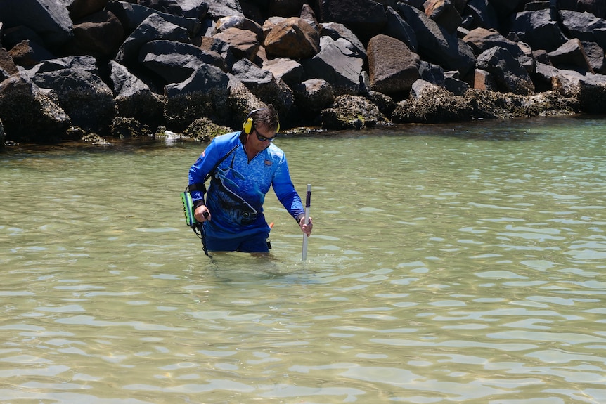 A man wades through shallow calm water with a metal detector and wearing headphones.