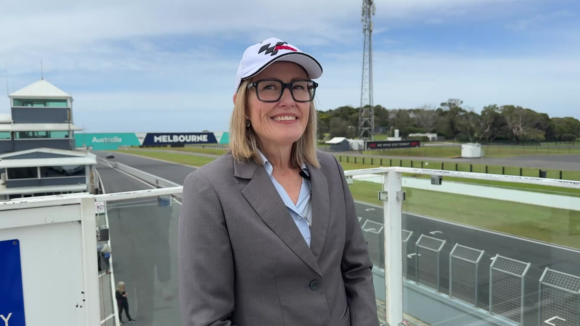 Uma mulher loira e de óculos está usando um boné de motoGP, parada ao lado da pista de corrida. 