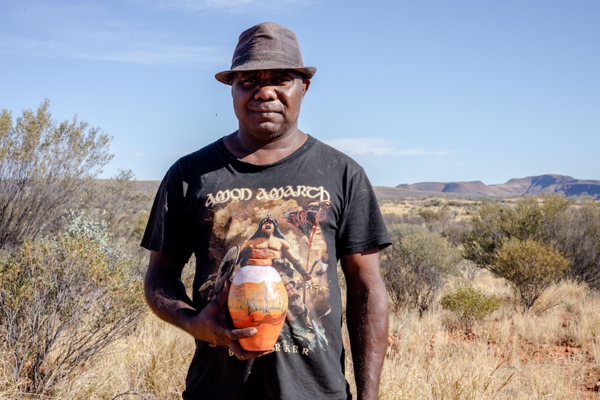 A man in a black t shirt and a trilby hat stands holding a brightly painted ceramic pot.