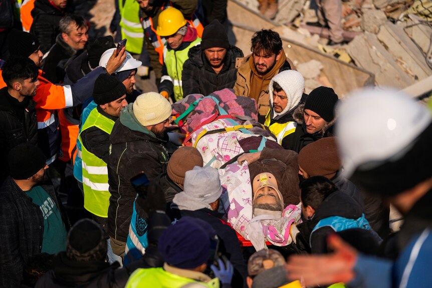 Elderly woman carried on stretcher by rescue workers through rubble of building.