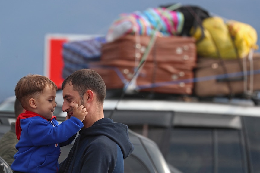 A man holding a toddler boy in front of a car with suitcases attached to the roof. 