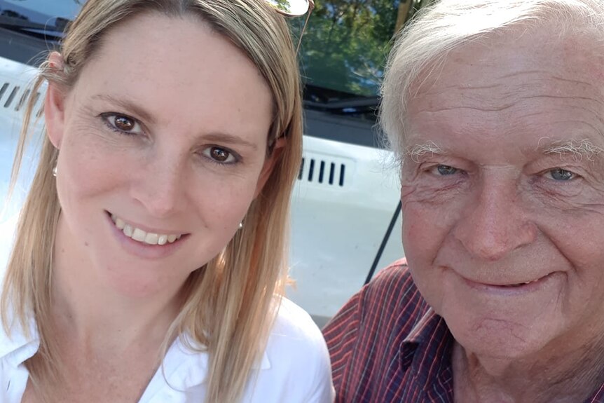 A blonde woman with her older white-haired father in front of a white vehicle.