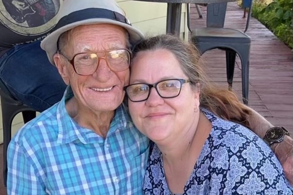An elderly man wearing glasses and a hat smiles for the camera with a woman with brown hair and glasses