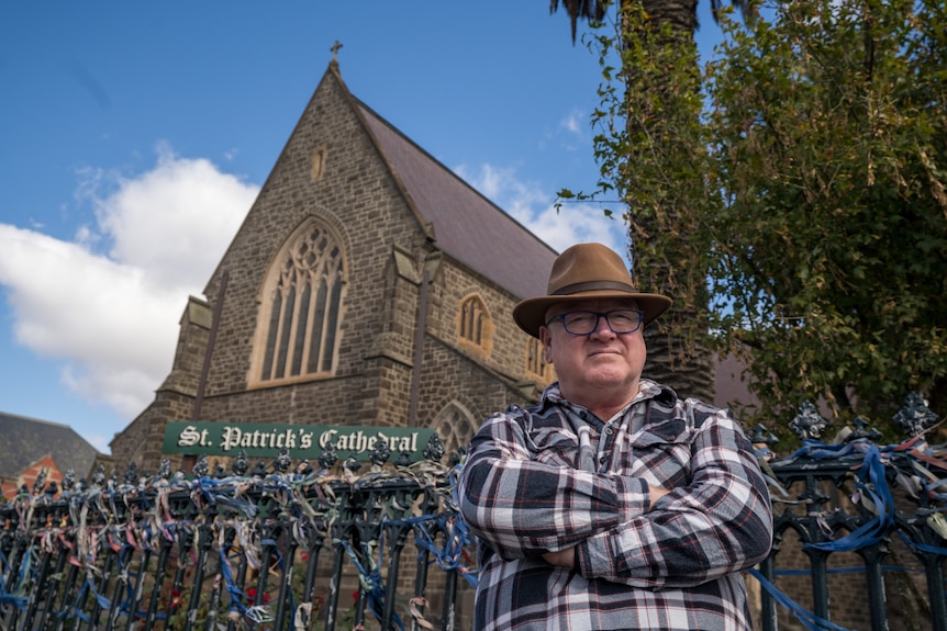 A man in a chequered shirt and hat stands outside a large stone church with ribbons on the fence.