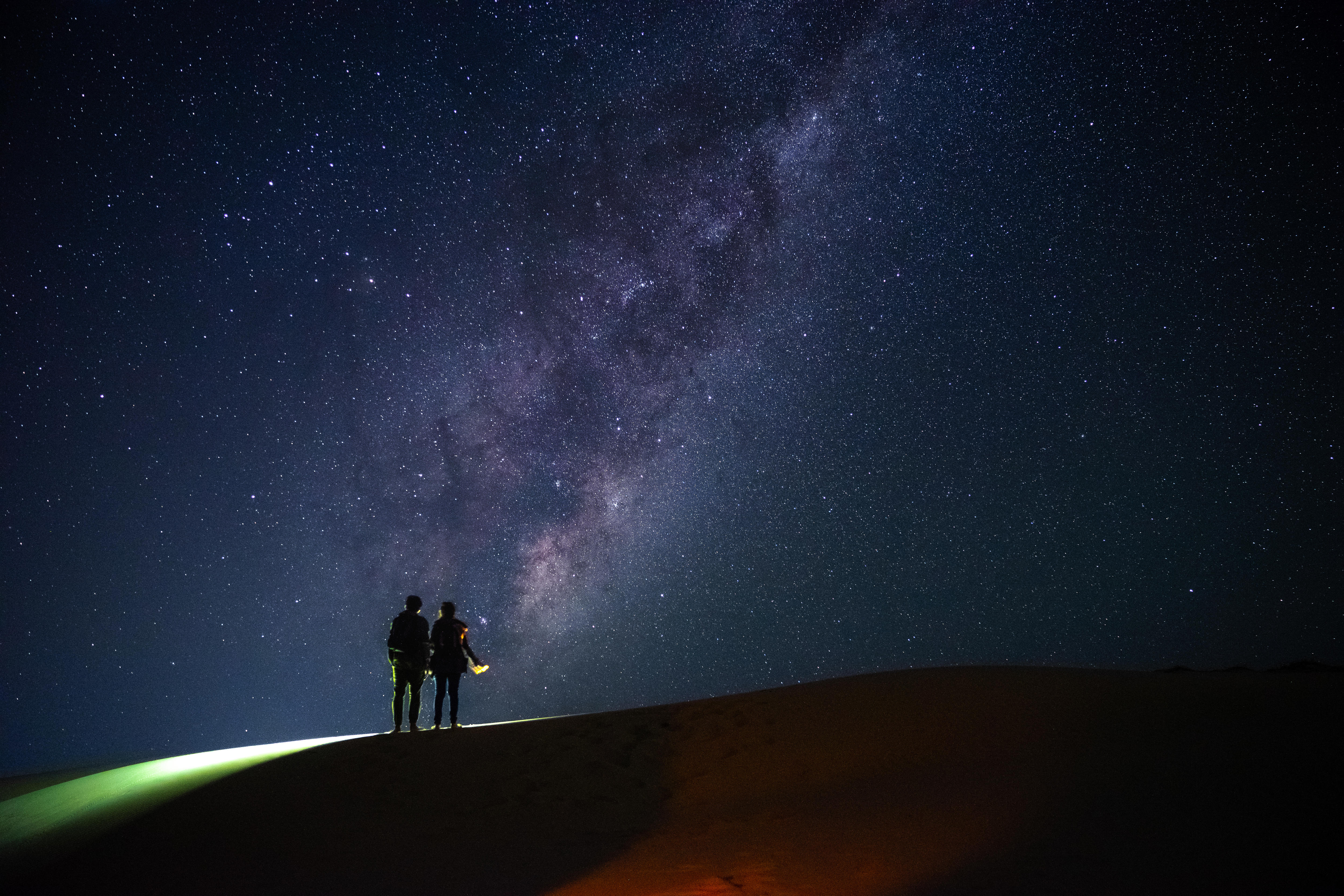 Two people are standing on a dark sand dune with the night sky behind lit up