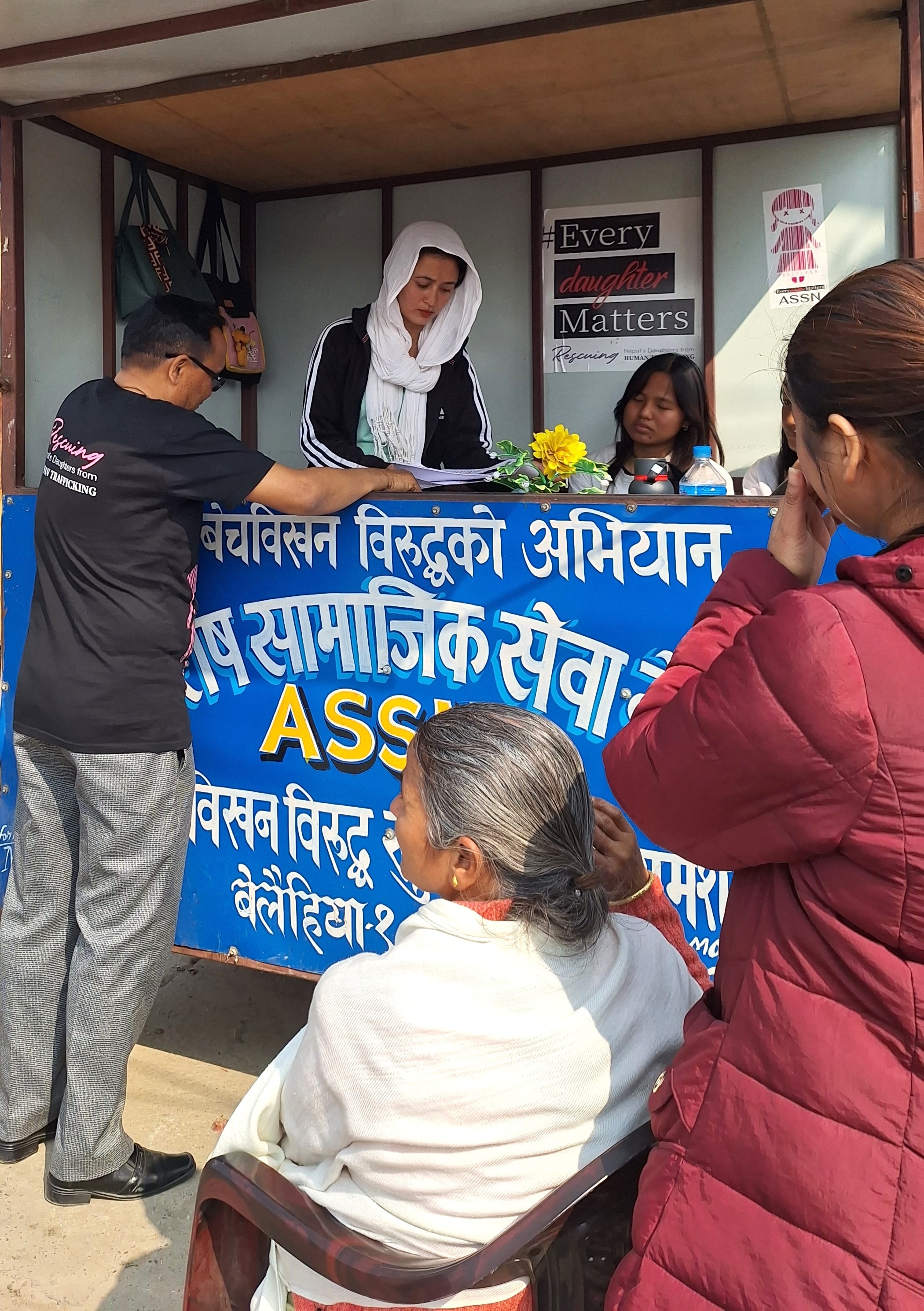 Uma cabine com duas mulheres conversando com pessoas do outro lado e algumas placas em nepalês e inglês.