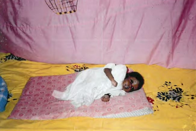 A baby in a white baby suit lies on a pink rug on top of a yellow rug.
