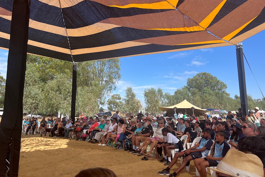 A group of indigenous people sit on chairs in an arc on the sand watching a performance