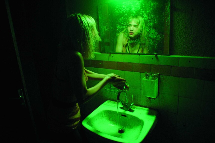 Colour still of Sofia Boutella looking into bathroom mirror under green light in 2018 film Climax.