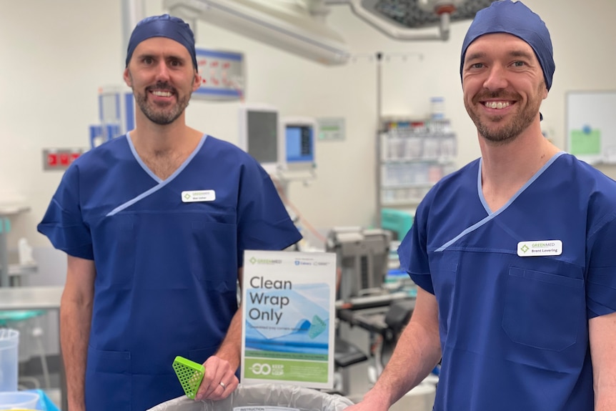 Two bearded men wearing dark blue scrubs smile for the camera in an operating theatre.