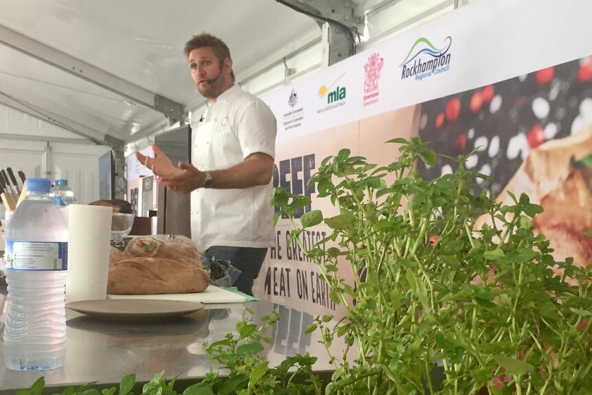 Curtis Stone stands in front of a bench speaking to a crowd.