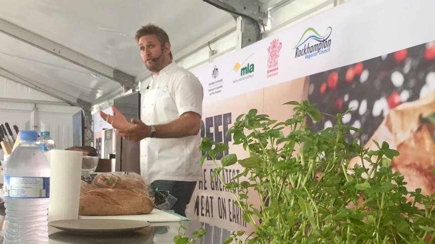 Curtis Stone stands in front of a bench speaking to a crowd.