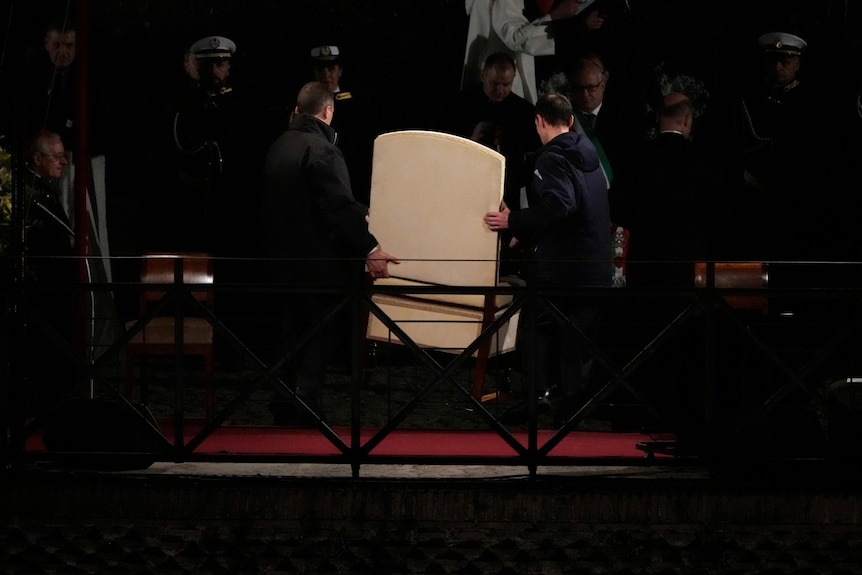 Two men in black clothing carry a large white chair off a podium.