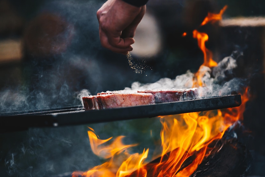 Steak is seasoned over an outdoor fire.