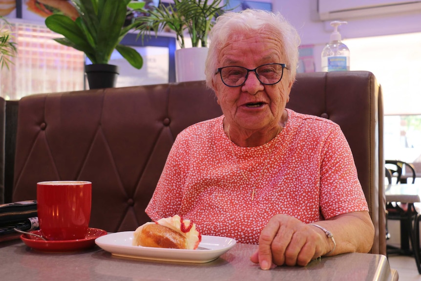 A woman wearing glasses sits in a cafe booth with a coffee cup and cake on the table in front of her.