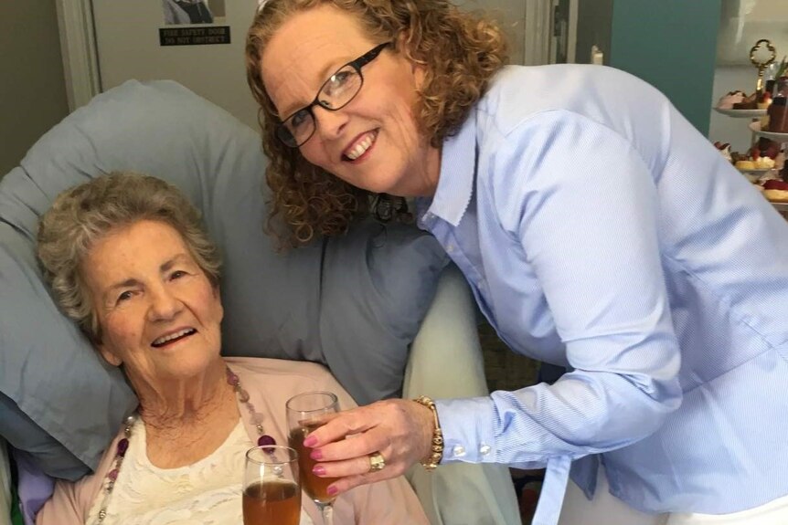 An elderly woman sits up in bed clinking wine glasses with a younger woman nearby.