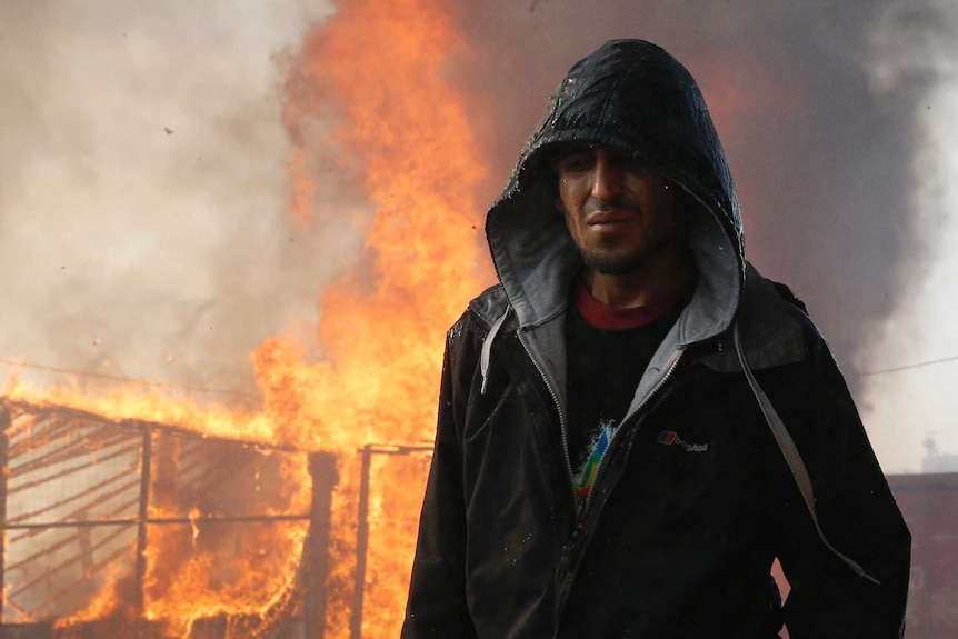 Man wearing hooded jacket walks past burning makeshift shelter
