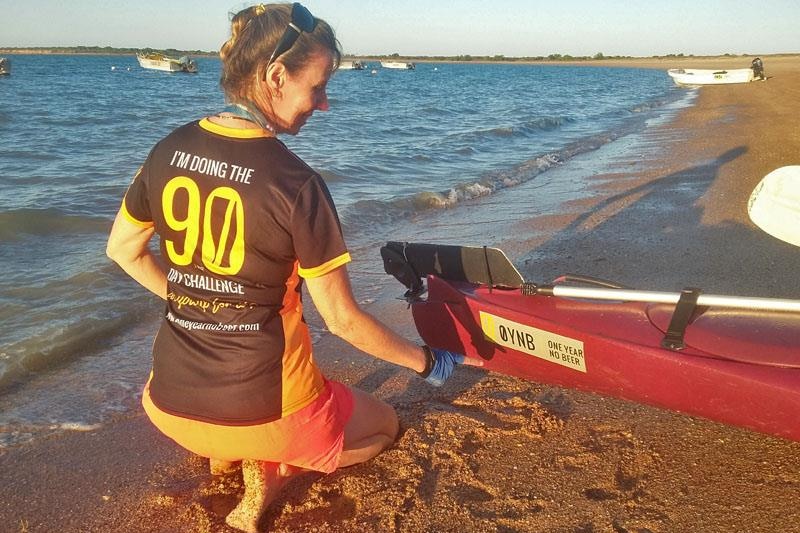 A woman kneels in the sand pointing at her kayak which has a 'one year no beer' challenge sticker on it