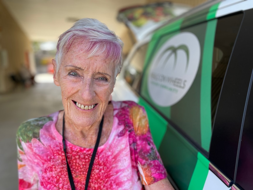 a woman in her seventies with grey and pink hair and a bright t shirt stands next to a meals on wheels van