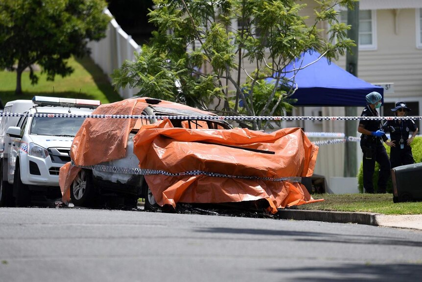 An orange tarp covers the burnt out car, which is surrounded by police tape.