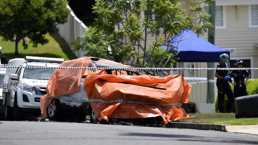 An orange tarp covers the burnt out car, which is surrounded by police tape.