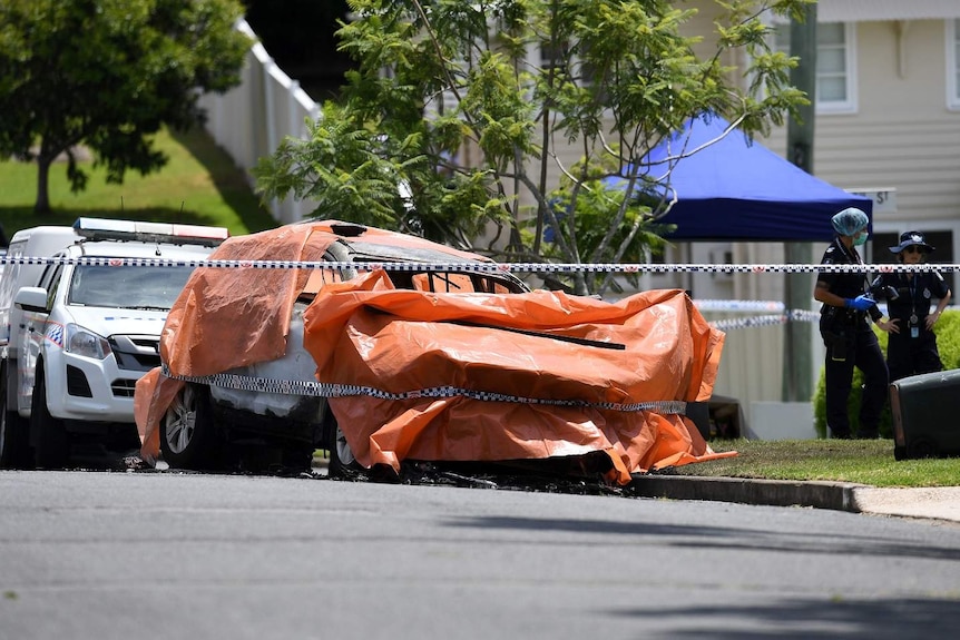 An orange tarp covers the burnt out car, which is surrounded by police tape.