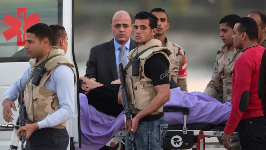Former Egyptian Prime Minister Hosni Mubarak lies in a stretcher as medical and security personnel escort him from a helicopter.