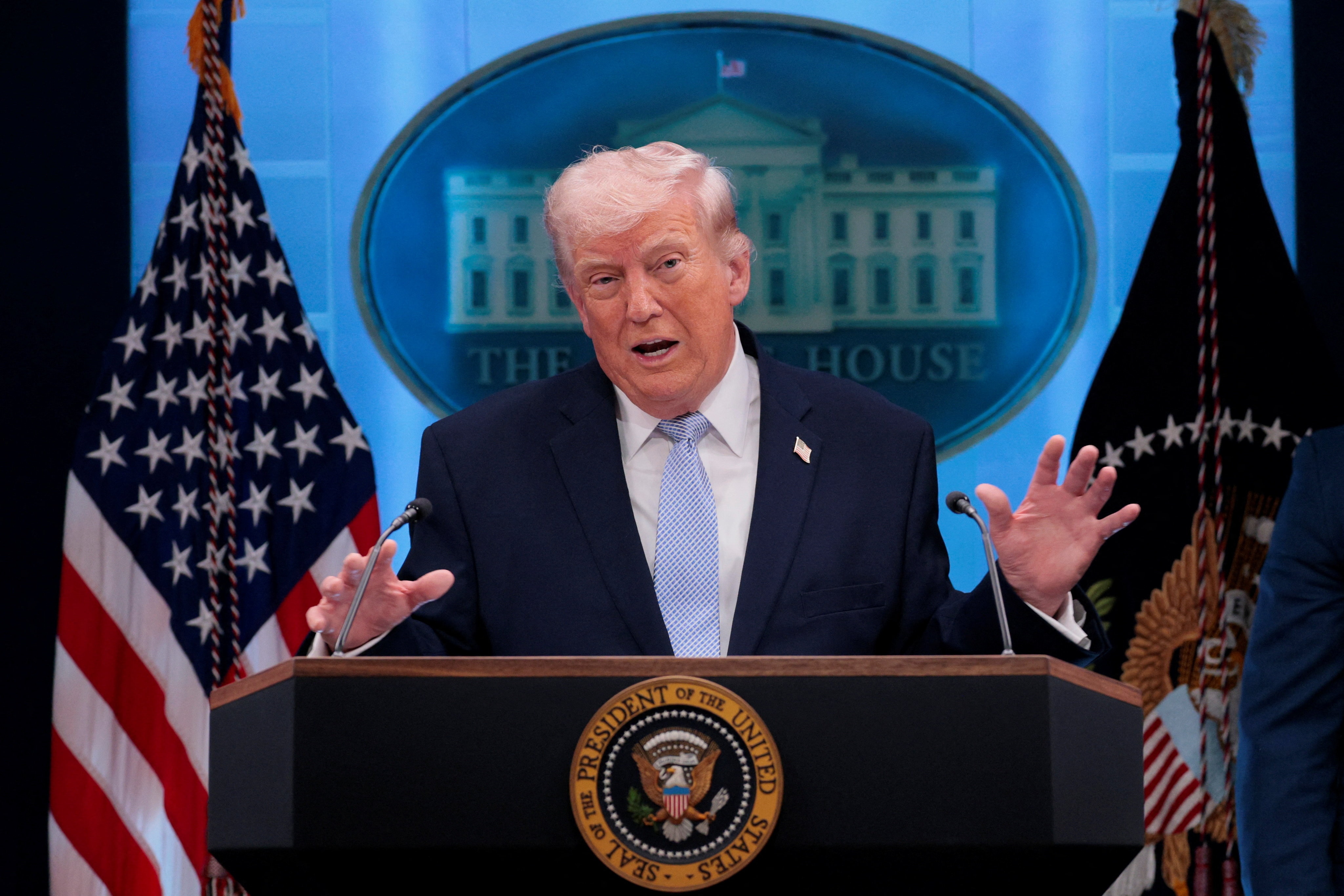 Donald Trump in a navy suit and lighter blue tie speaking at a lectern with both hands raised, in front of a White House sign.