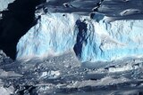 An aerial view of a bluish-white giant glacier with craggy sides and a snowswept top