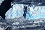 An aerial view of a bluish-white giant glacier with craggy sides and a snowswept top