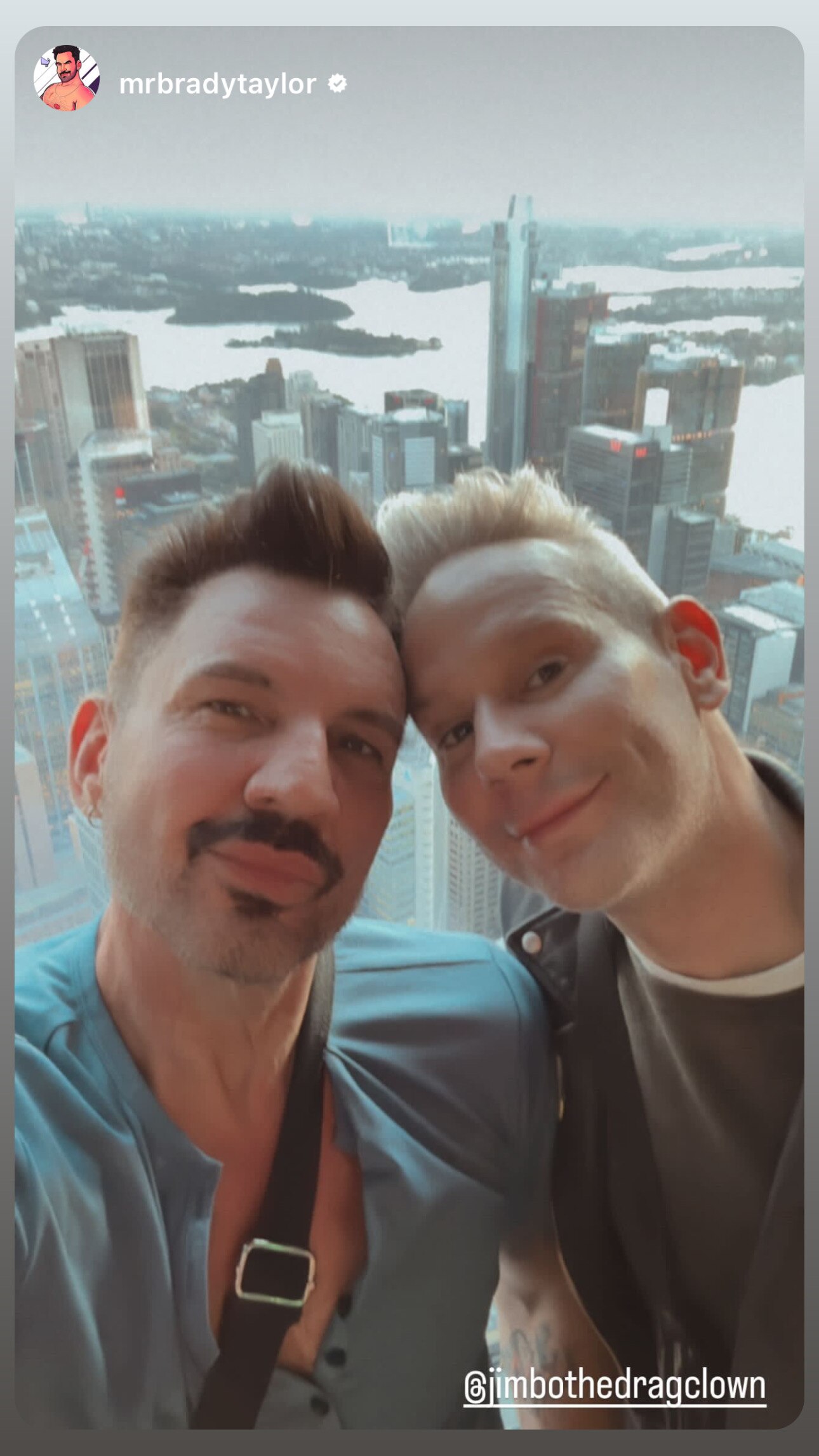 Two men smile for a selfie, heads together. A window reveals Sydney's skiline, the two high above.