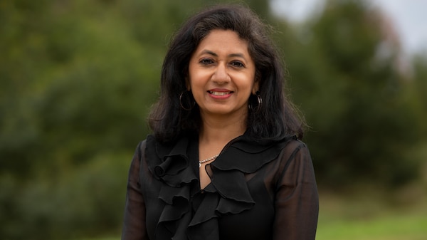 Portrait of a middle-aged Indian woman outside surrounded by greenery. She is wearing a black top and has shoulder length hair.