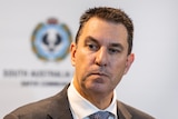 A man with a serious expression wearing a suit and tie stands in front of an SA Police banner