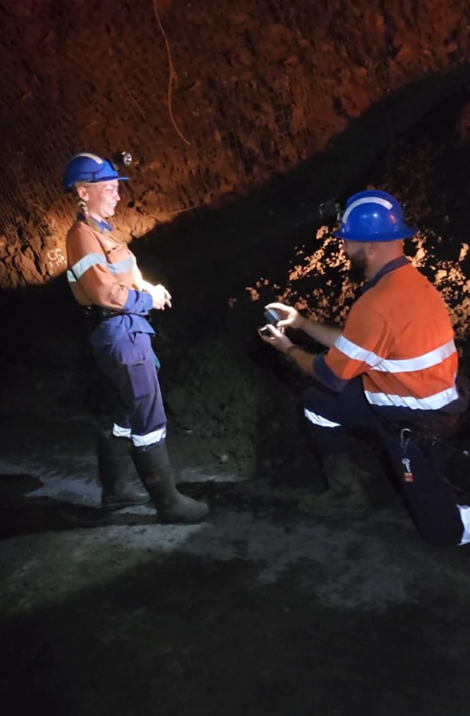 A man in orange bends down on one knee with a ring box in hand to his smiling partner