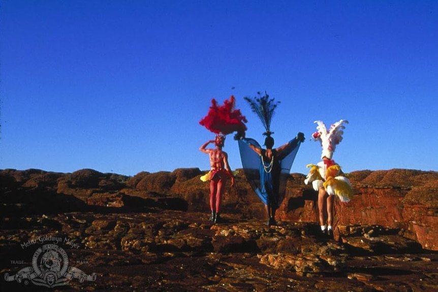 Three drag queens stand in front of an outback Australian canyon wearing large headpieces and colourful costumes