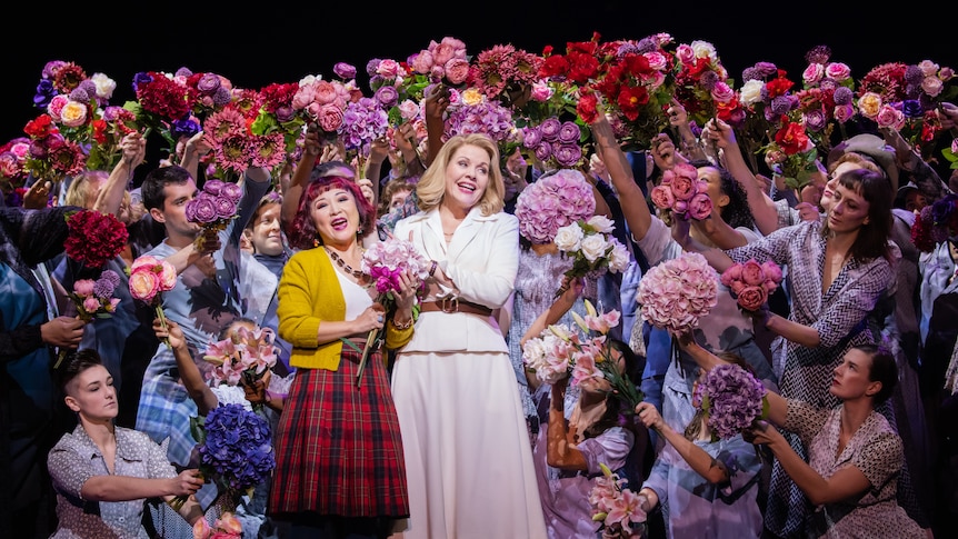 Two women sing while smiling in front of a chorus who surround them with bunches of flowers