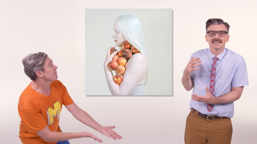 Two men stand  either side of a painting of a woman holding fruit
