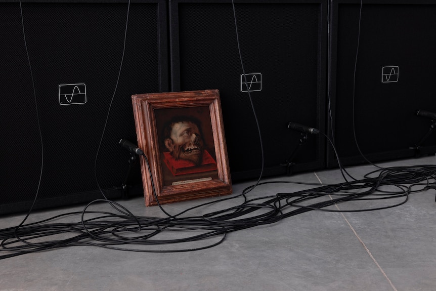 A framed medieval picture of a decapitated head on a book rests against a row of amps and electrical cords
