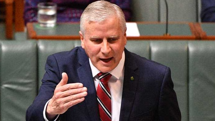 A man wearing a navy suit and red, white and blue striped tie gestures and talks with green leather seats behind him