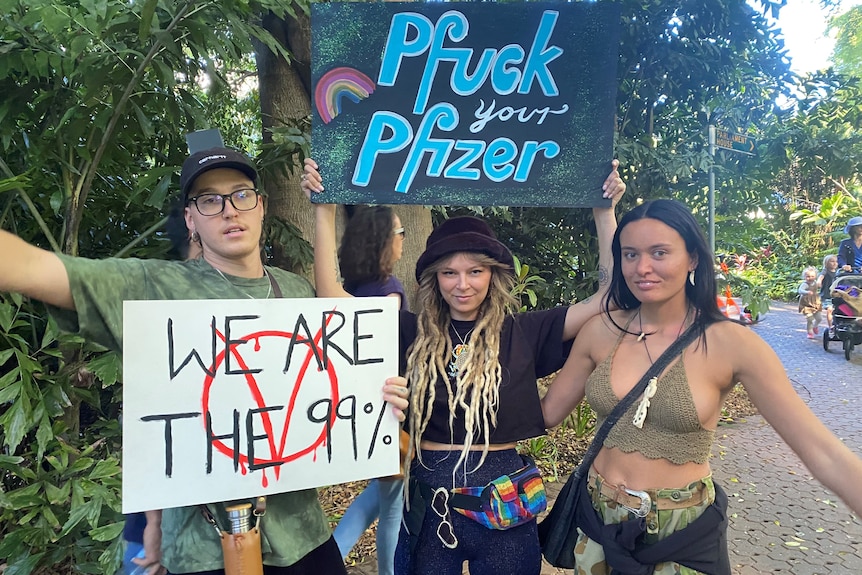 Three young people with pierced septums and dreadlocks, holding signs saying "we are the 99 per cent" and "pfuck your Pfizer". 
