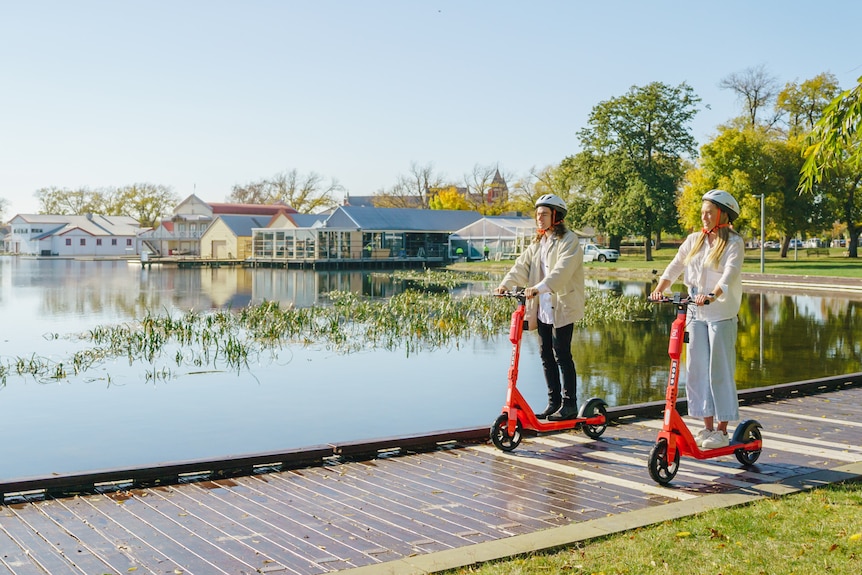 Two people smile and ride scooters along the boardwalk of a lake