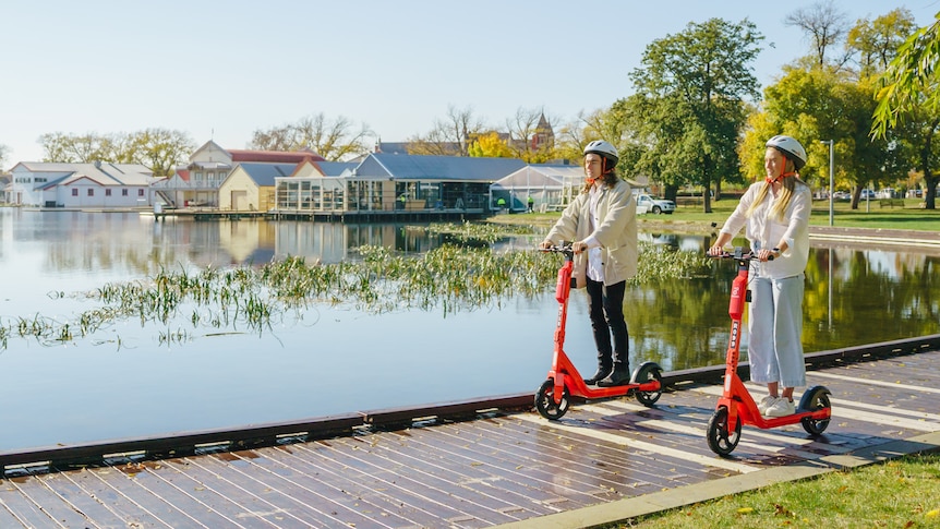 Two people smile and ride scooters along the boardwalk of a lake.